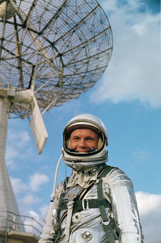 Wearing his silver Mercury pressure suit, John Glenn is shown smiling in front of a large telescope array at Cape Canaveral