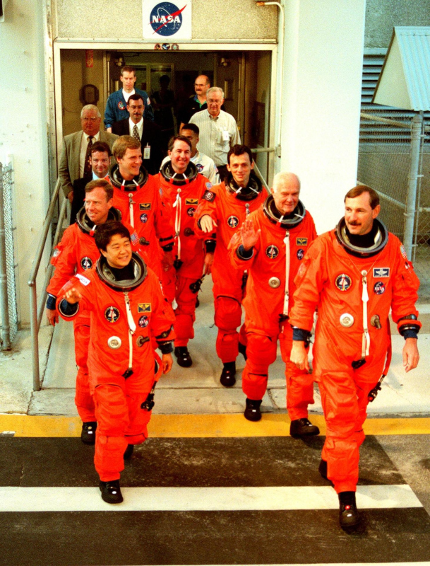 The crew of STS-95, dressed in their orange launch suits, walk from the Operations and Checkout Building on their way to the launch pad